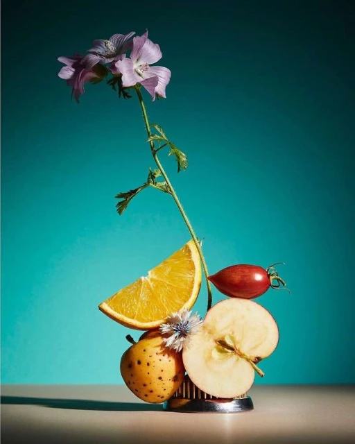 Still life photography featuring a vibrant arrangement of flowers, fruits, and botanical elements against a bright teal backdrop, creating a colourful, organic composition.