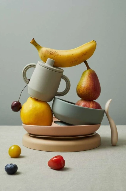 Still life of a banana, pear, cherry, lemon, apple, and dishes stacked on a table against a solid gray background.