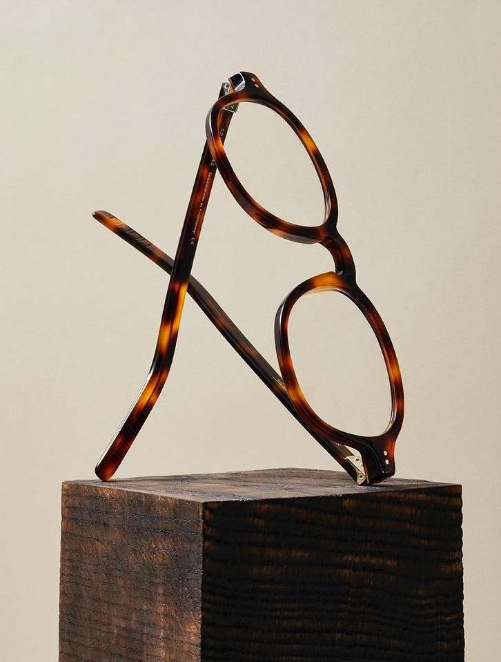 Tortoiseshell eyeglasses balanced on a dark, stained wood block against a soft neutral backdrop, showcasing eyewear design.