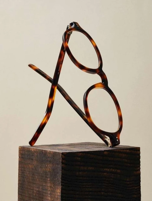 Tortoiseshell eyeglasses balanced on a dark, stained wood block against a soft neutral backdrop, showcasing eyewear design.