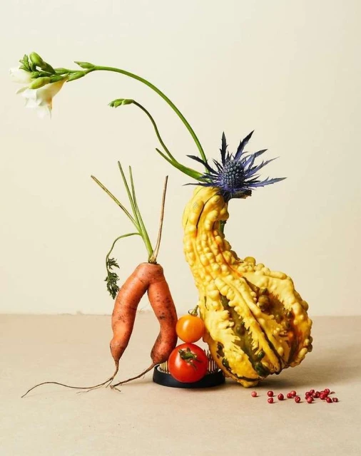 Still life arrangement of gourds, carrots, tomatoes, and flowers in a neutral-toned indoor setting, showcasing a natural and artistic composition.