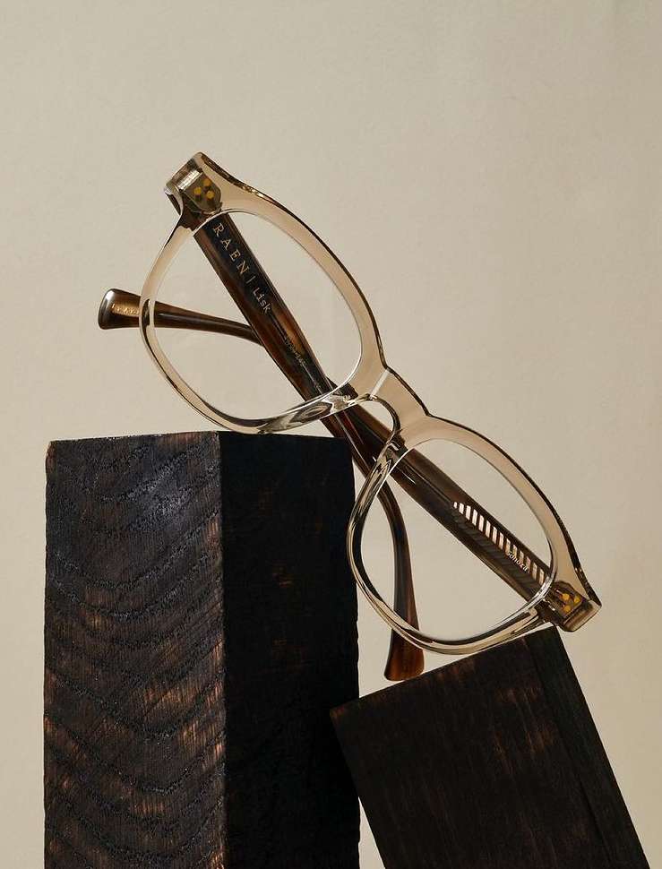 Close-up of stylish eyewear glasses by Raen, featuring clear lenses and a neutral frame, placed on rustic wood blocks.