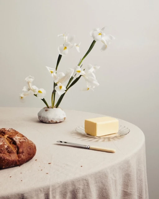 Still life of loaf of bread, butter on a plate, knife, and white flowers in a vase, set on a round table with a white tablecloth.