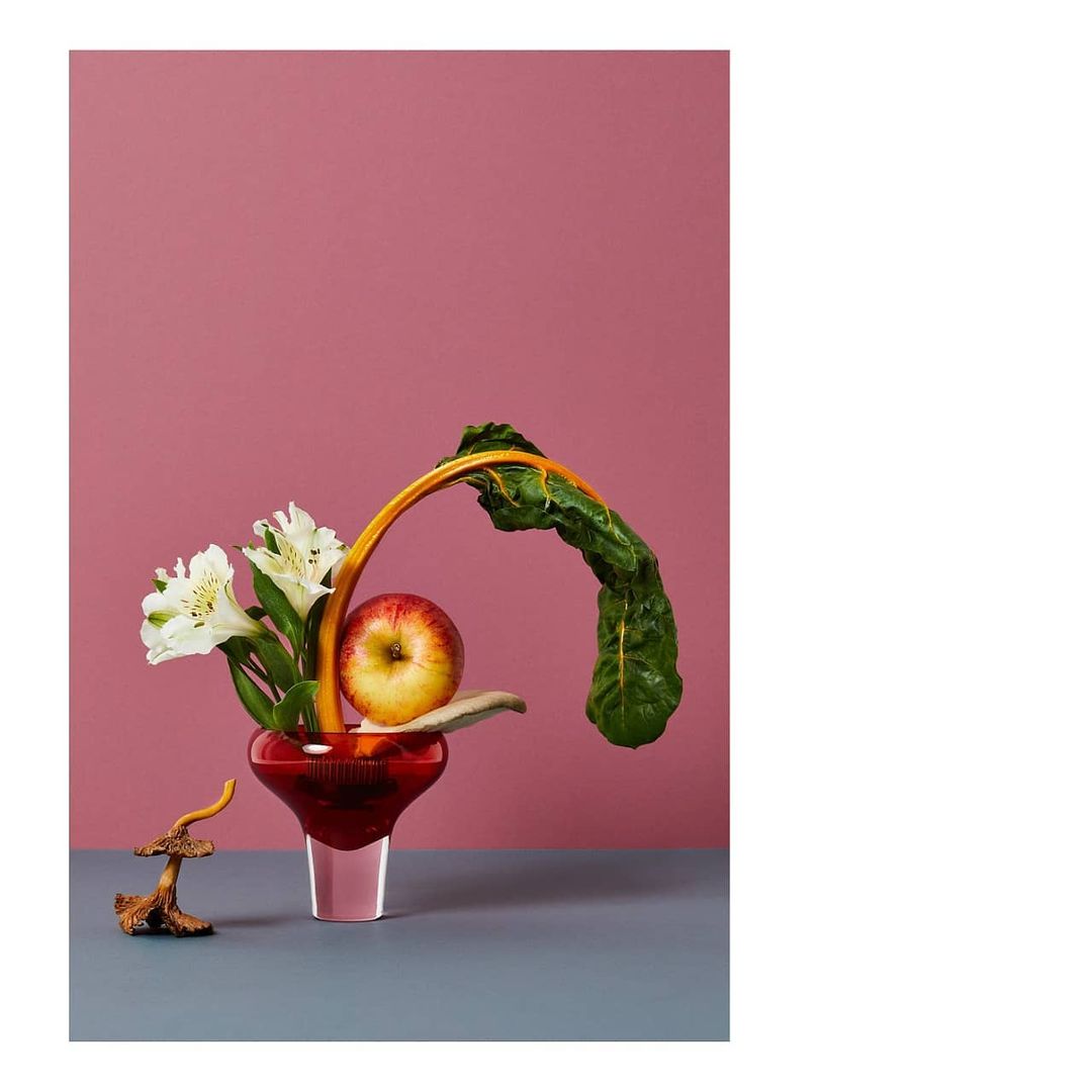 A still life photograph features white flowers, an apple, and vibrant chard leaves artfully arranged in a red glass vase against a mauve backdrop.