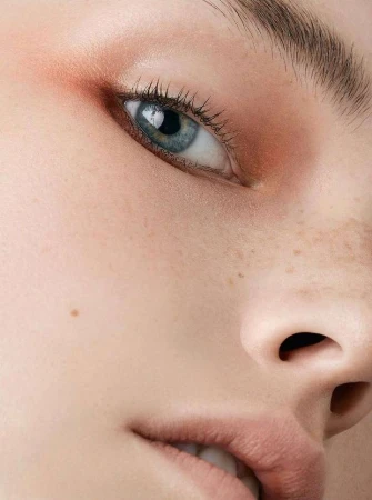 Close-up studio shot of a young adult female's face, showcasing her blue eye, light skin, freckles, and makeup with warm, neutral tones.
