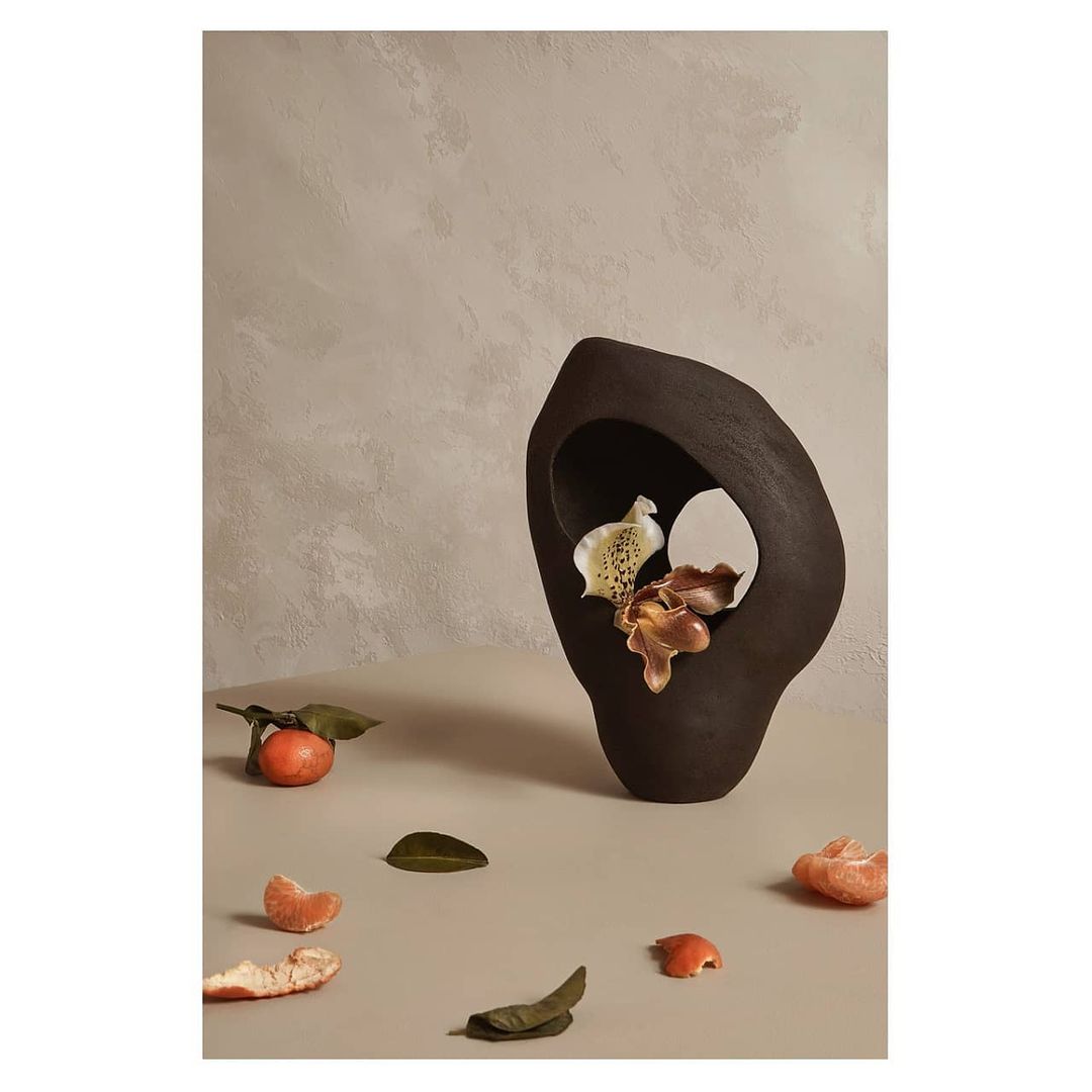 Still life shot of a dark brown ceramic vase holding flowers with oranges and orange peels scattered across a beige table surface.
