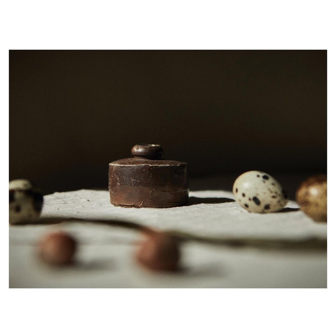 Close-up still life of a brown ceramic lidded jar surrounded by speckled eggs, sitting on textured paper under soft, diffused light.