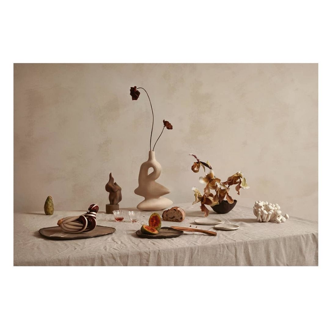 Still life on a draped linen cloth features a modern vase with dried floral elements, pottery, cut fruit, and decorative coral.
