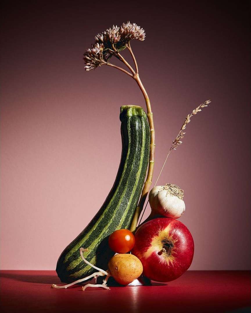 Still life photograph of zucchini, apple, tomato, garlic, potato, and flowers against a gradient pink background.