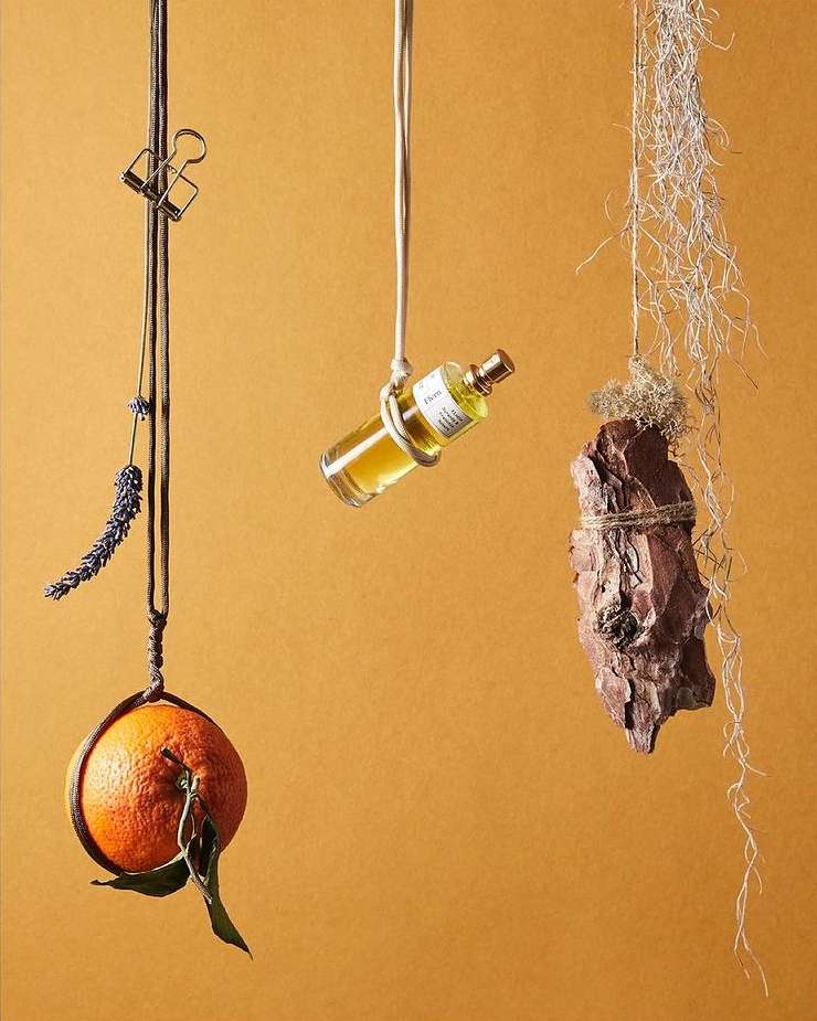 Still life featuring an orange, lavender, perfume bottle, and tree bark suspended by strings against an orange-yellow background.