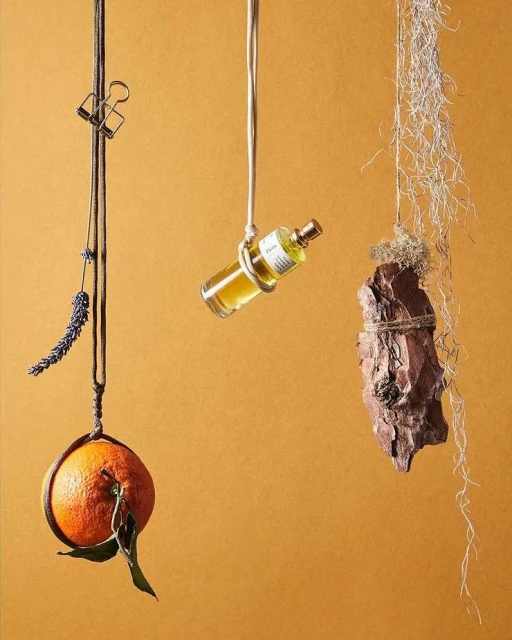 Still life featuring an orange, lavender, perfume bottle, and tree bark suspended by strings against an orange-yellow background.
