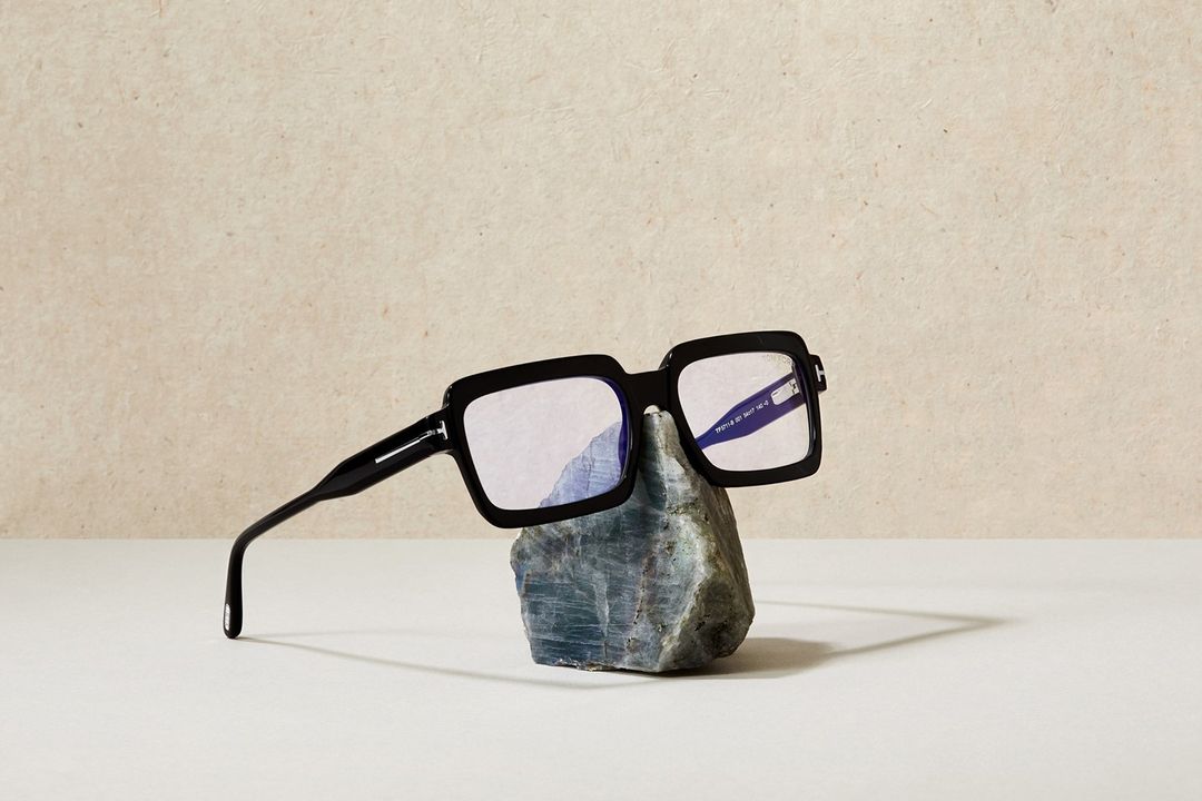 Black square eyeglasses sit on a grey rock in a minimalist, neutral-toned product shot. The background is a textured beige.