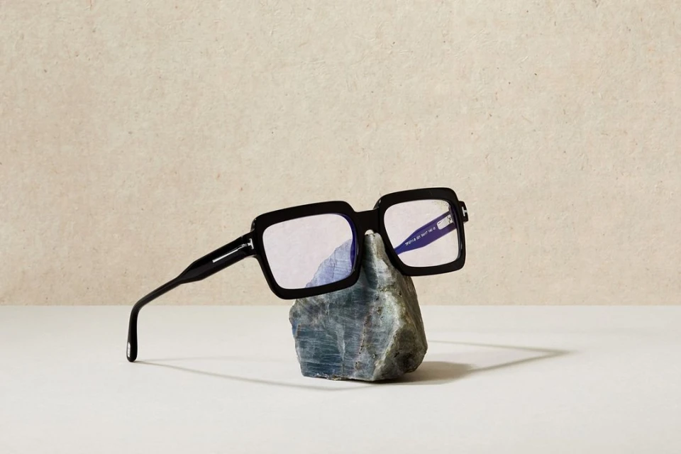 Black square eyeglasses sit on a grey rock in a minimalist, neutral-toned product shot. The background is a textured beige.