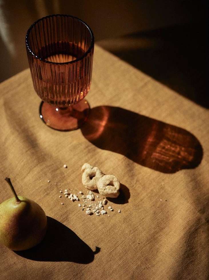 Still life featuring a ribbed amber glass filled with liquid, alongside a pear and two small ring-shaped crackers on a textured fabric surface.