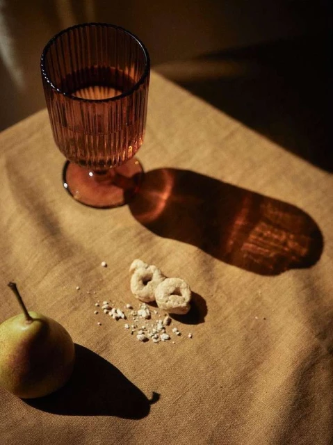 Still life featuring a ribbed amber glass filled with liquid, alongside a pear and two small ring-shaped crackers on a textured fabric surface.