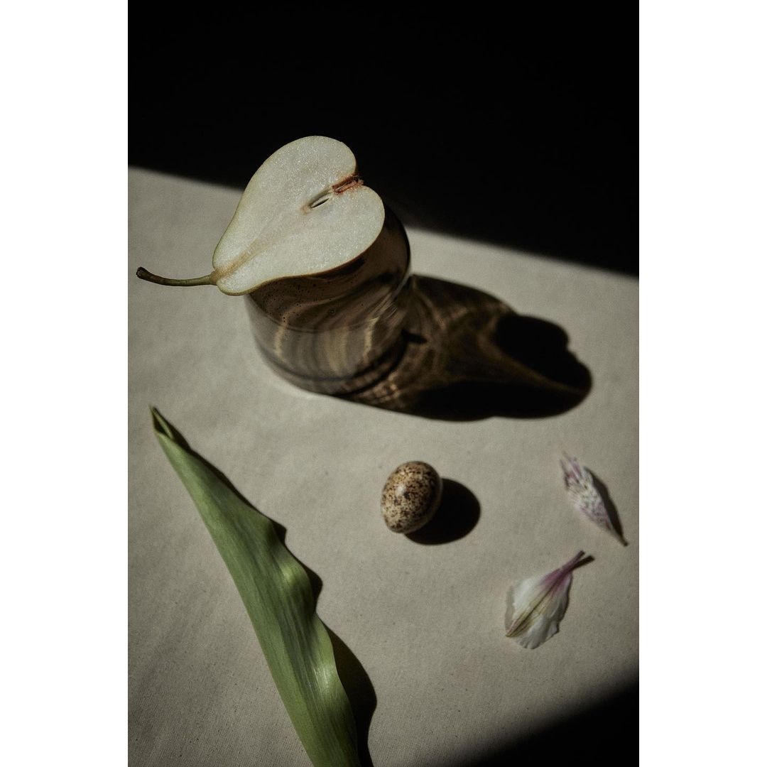 Still life photograph of a halved pear with a green stem, a quail egg, and a flower petal on a beige surface.