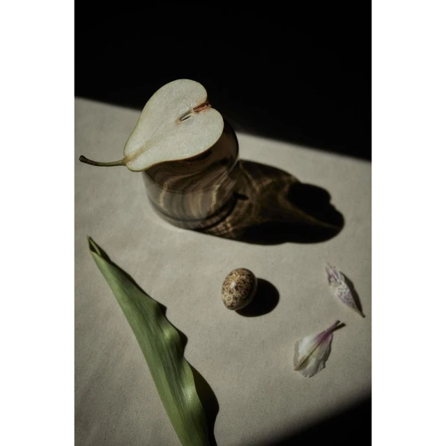 Still life photograph of a halved pear with a green stem, a quail egg, and a flower petal on a beige surface.