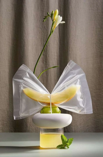 Still life shot of melon slice held in a plastic wrap bow, set on a lime, sitting atop a white plinth, with yellow liquid in glass below.