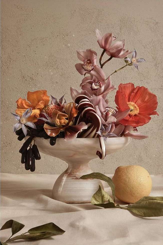 Still life photography of a floral arrangement in a white marble bowl next to a lemon, set against a beige textured background.