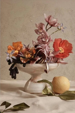 Still life photography of a floral arrangement in a white marble bowl next to a lemon, set against a beige textured background.