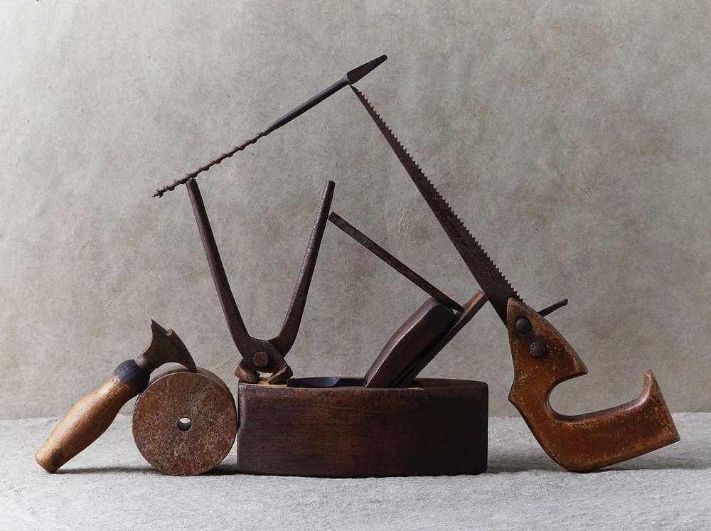 Still life shot of vintage hand tools including saws, metal cutters, and a wooden block plane on a neutral surface.