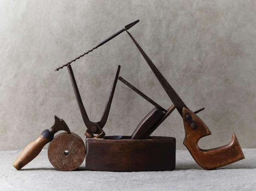Still life shot of vintage hand tools including saws, metal cutters, and a wooden block plane on a neutral surface.