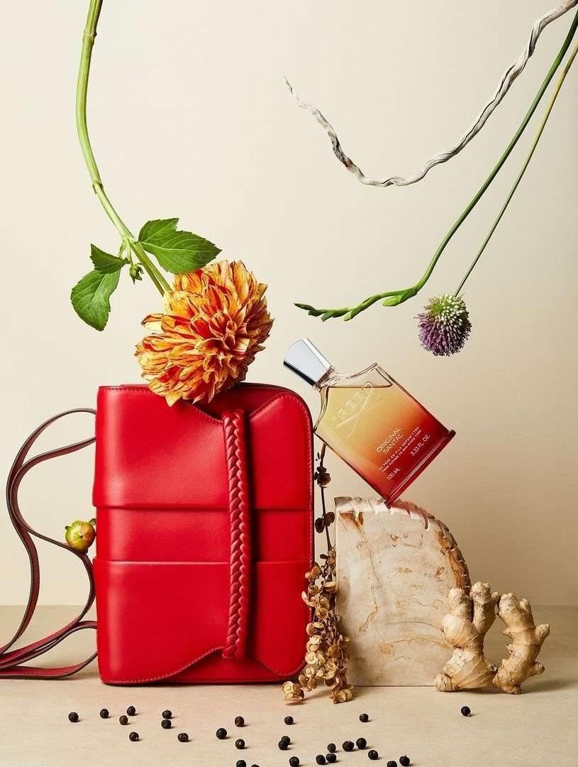 Still life shot of a red leather handbag, a glass perfume bottle, an orange flower, ginger root, and black peppercorns arranged on a light background.