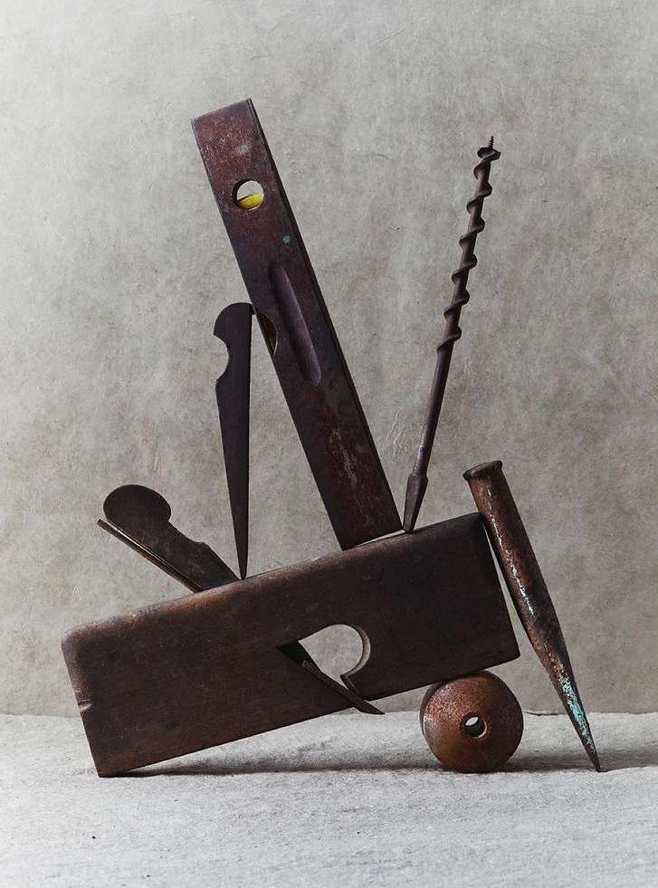 Still life photo of old, rusty hand tools including a plane, level, nail, and drill bit arranged on a neutral-colored surface.