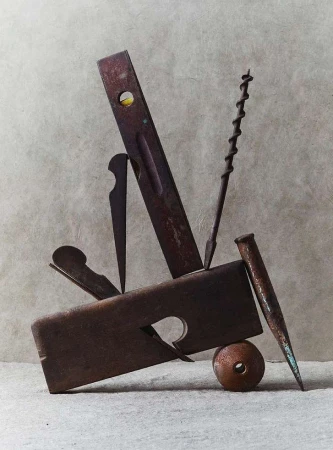Still life photo of old, rusty hand tools including a plane, level, nail, and drill bit arranged on a neutral-colored surface.