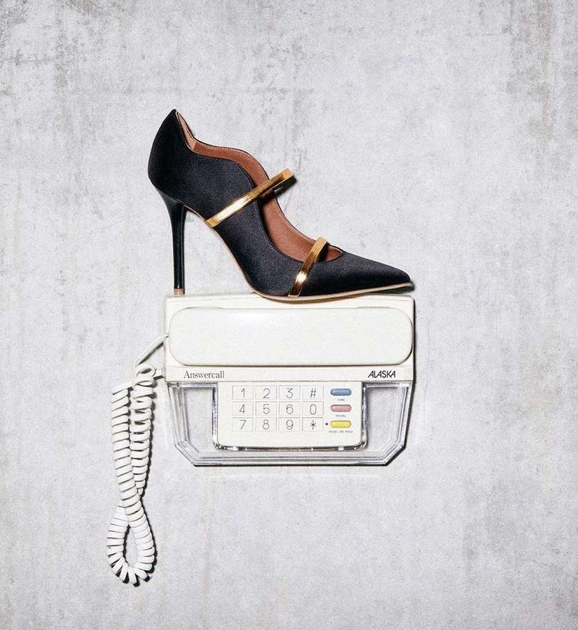 Black high-heel shoe with gold trim sitting on a vintage white telephone against a grey textured wall in a studio setting.
