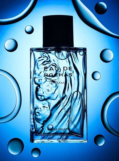 Studio shot of a clear glass Eau de Rochas Homme fragrance bottle with black cap against a blue background with floating translucent bubbles.