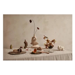 Still life table setting featuring an abstract ceramic vase, dried flowers, fruit, and pottery on a textured linen tablecloth.