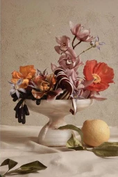 Still life arrangement of flowers, fruits, and a red poppy in a marble pedestal bowl with a yellow lemon resting on fabric below.