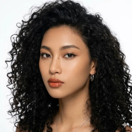 Close-up portrait of a woman with voluminous curly black hair, glowing makeup, and glossy lips against a white background.
