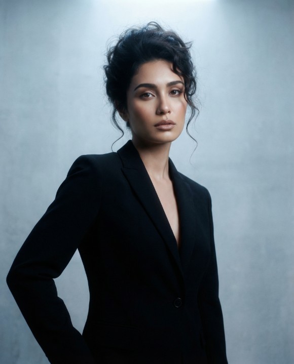 A woman with a dark curly updo wearing a tailored black blazer against a cool grey-blue textured backdrop.