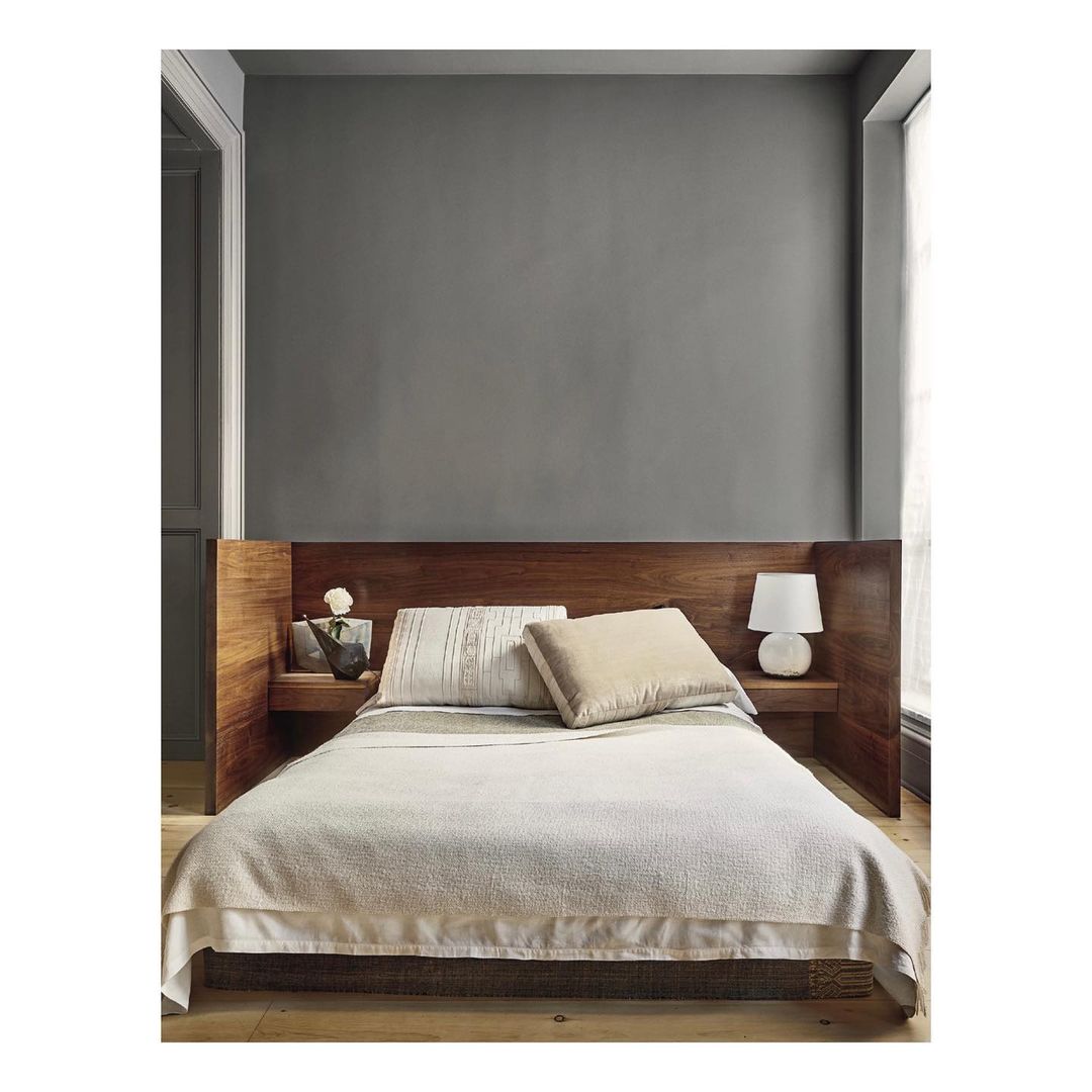 Bedroom interior featuring a queen-size bed with a wooden headboard, white bedding, side table with lamp and decorative items, neutral color palette.