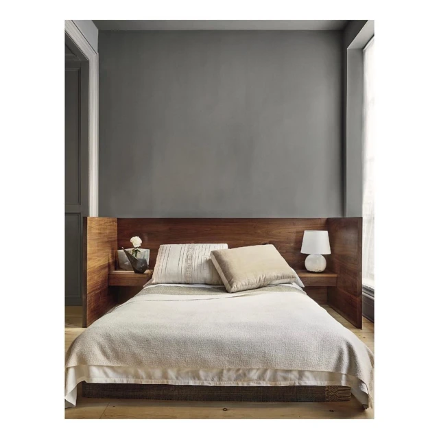 Bedroom interior featuring a queen-size bed with a wooden headboard, white bedding, side table with lamp and decorative items, neutral color palette.
