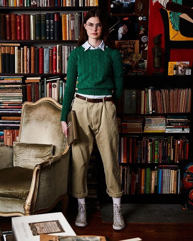 Young woman in glasses stands amid bookshelves wearing a green sweater and tan pants, holding a book in front of a velvet chair.