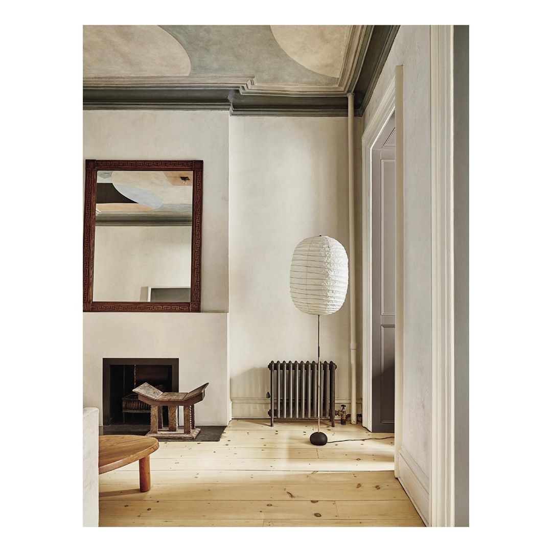 Interior space featuring a floor lamp with a paper shade, a fireplace with a mirror above, a wooden stool, and light wood flooring.