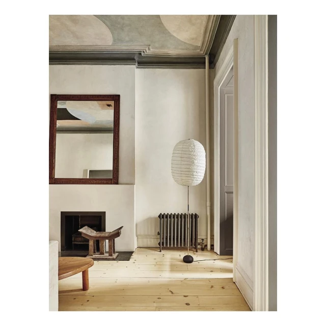 Interior space featuring a floor lamp with a paper shade, a fireplace with a mirror above, a wooden stool, and light wood flooring.