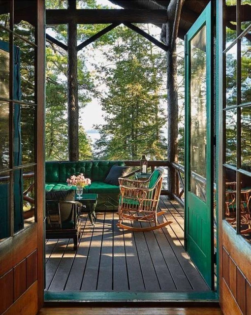 Veranda scene with green upholstered couch, rocking chair, and coffee table, set against a backdrop of lush trees.