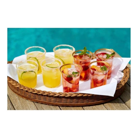 Eight glasses of colorful cocktails with fruit and mint sit on a woven tray atop a wooden surface near a blue swimming pool.