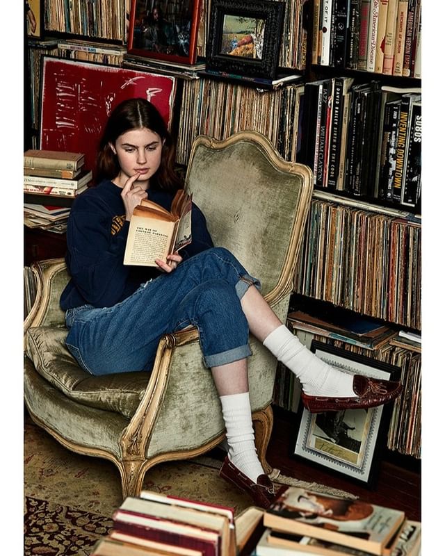 Young woman sits in an armchair, absorbed in reading a book, surrounded by bookshelves filled with books and vinyl records in a room.