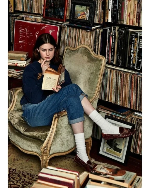 Young woman sits in an armchair, absorbed in reading a book, surrounded by bookshelves filled with books and vinyl records in a room.