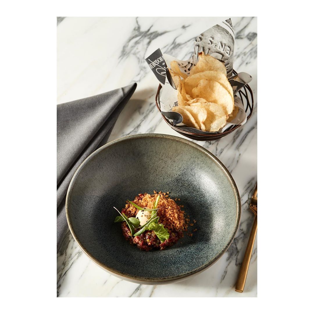 Bowl with a small serving of beef tartare topped with microgreens, served with a basket of potato chips on a marble table.
