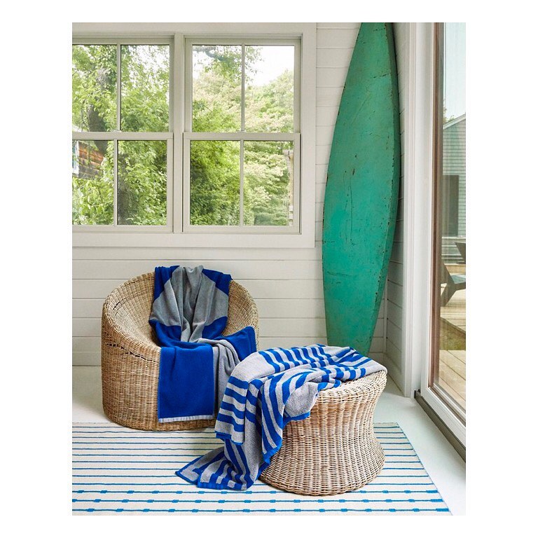 Indoor lounge area featuring a rattan chair and ottoman topped with blue and grey striped throws next to a decorative surfboard.