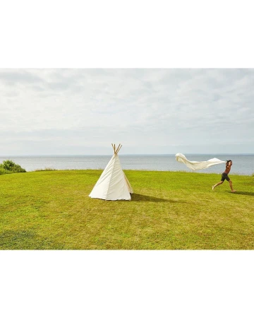 A person runs with a white fabric on a green lawn by a teepee near the ocean under a cloudy sky in a wide outdoor shot.