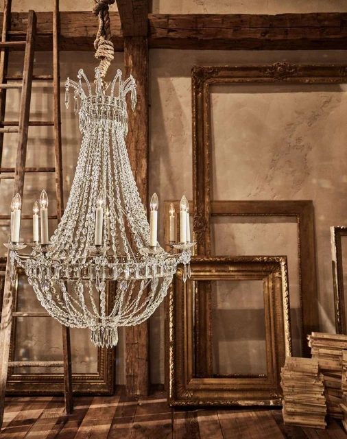 Crystal chandelier hanging in a rustic room with wooden floors and beams, complemented by empty picture frames and stacked books.