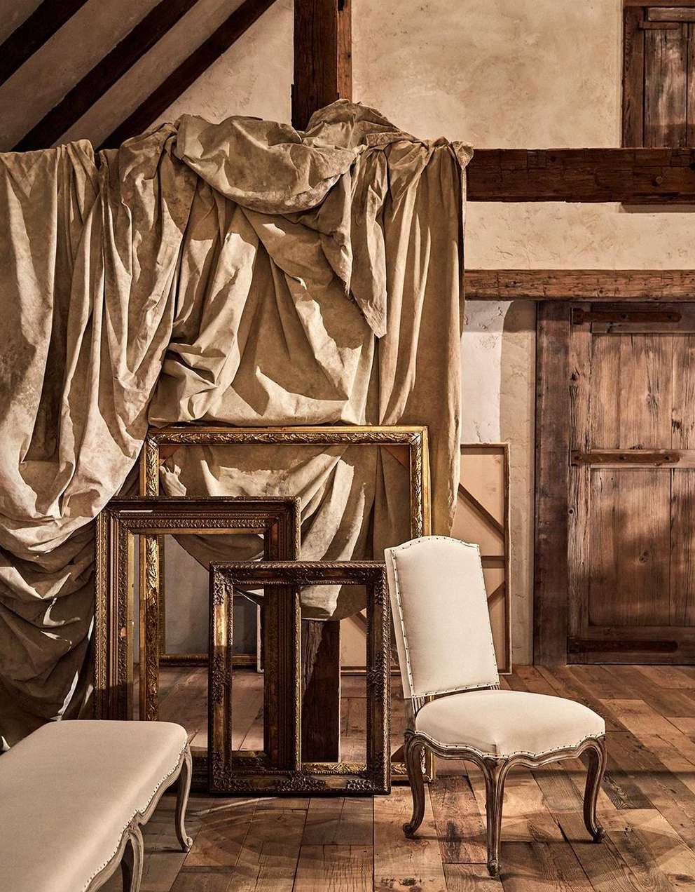 Interior composition with antique chair, draped textile, ornate empty picture frames, and wooden door, creating a vintage aesthetic.
