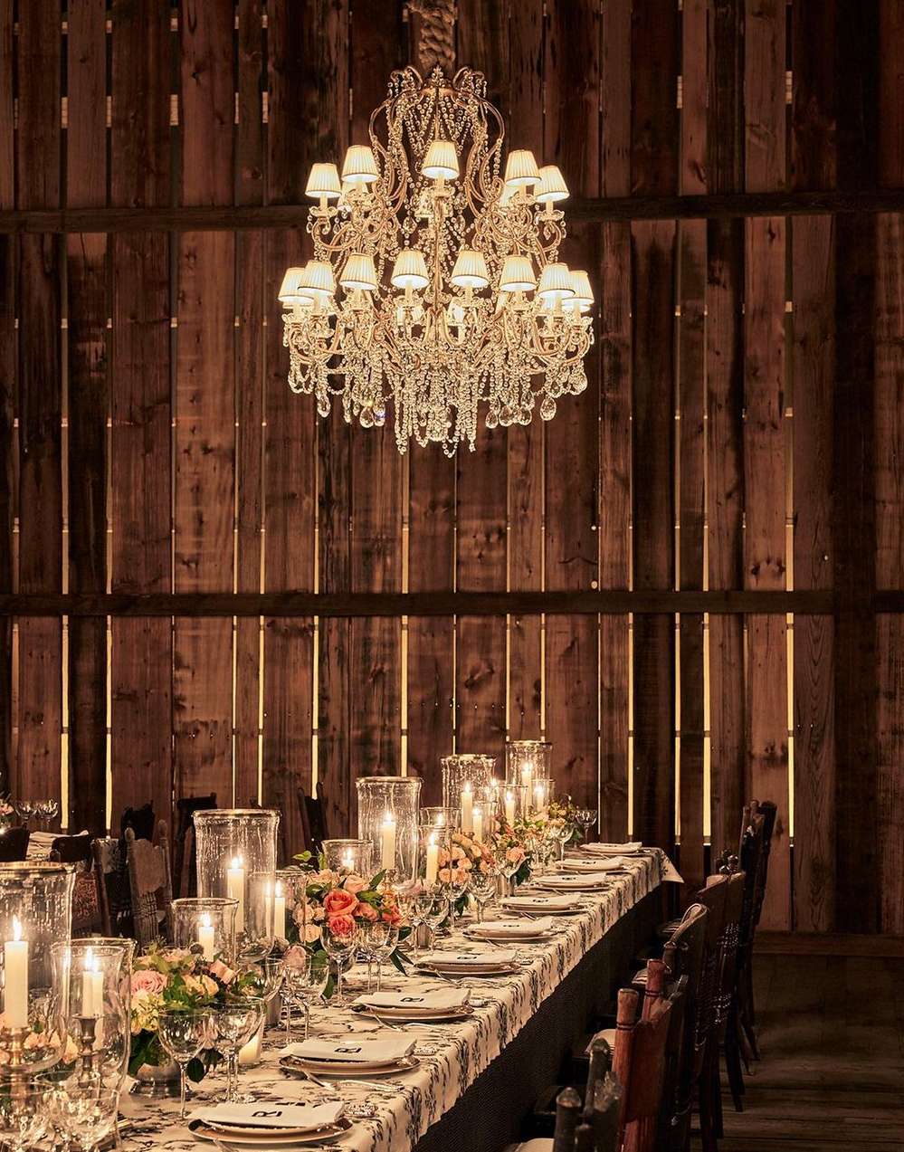 Crystal chandelier illuminates a long formal dining table set for an event in a barn interior with rustic wood walls and flooring.